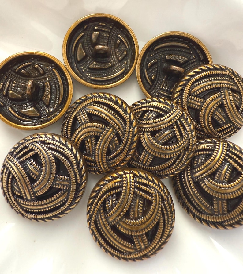 May include: A collection of antique brass buttons with intricate, swirling designs. The buttons have a dark, textured background with raised, golden-colored detailing. The buttons are round and vary in size.