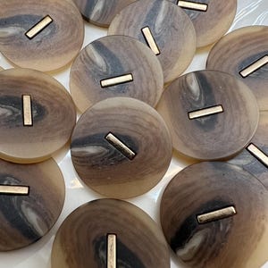 May include: A collection of round buttons with a marbled design in shades of brown, tan, and gray. Each button features a rectangular gold-colored metal insert. The buttons are arranged on a white surface.