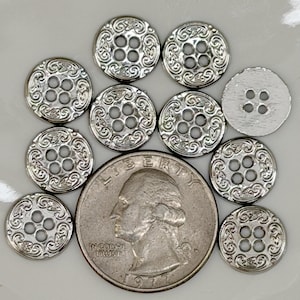 May include: A selection of silver-coloured buttons featuring an ornate, swirling pattern. Each button has four holes for sewing. A U.S. quarter is included for size comparison. The buttons are round and have a vintage aesthetic.