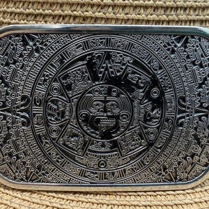 May include: A rectangular silver-toned belt buckle featuring a detailed, engraved design of the Mayan calendar. The intricate pattern includes circular and geometric shapes, with a central sun-like figure. The buckle is set against a straw hat.