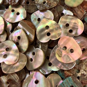 May include: A collection of heart-shaped buttons crafted from iridescent mother-of-pearl shell. Each button features two central holes for attachment and displays a unique, natural color variation ranging from pearlescent white to deep brown.