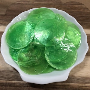 May include: A collection of circular, translucent green shell discs with a smooth, iridescent finish. Each flat disc features a small, centered hole near the top edge, designed for threading or attachment in various craft projects.