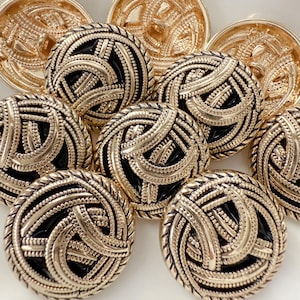 May include: Close-up of several gold-colored buttons with a black center. The buttons have a detailed, woven design around the edges and a circular pattern in the center. The buttons are arranged on a white surface.