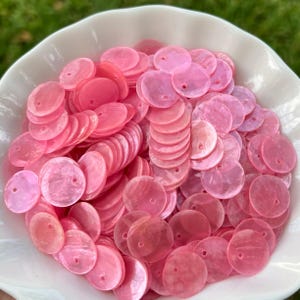May include: A collection of small, round, pink mother-of-pearl sequins with a single hole in the centre of each piece. These flat, circular discs feature a smooth, iridescent finish and are piled together in a white, scalloped-edge bowl.