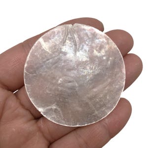 Puede incluir: Un disco de concha circular, translúcido e iridiscente con una superficie lisa y plana. El material muestra un brillo nacarado con variaciones sutiles en el reflejo de la luz, presentando una textura orgánica natural y una apariencia fina y semitransparente.