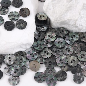 May include: A collection of small, round, dark gray buttons with a scalloped edge and a two-hole center. Each button features a subtle, iridescent sheen and a carved floral pattern on the surface, scattered across a white textured base.
