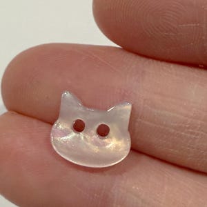 May include: A small, iridescent cat-shaped button with two dark brown eye holes. The button is pearlescent white with hints of pink and yellow, and has two small pointed ears.