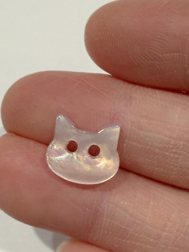 May include: A small, iridescent cat-shaped button with two dark brown eye holes. The button is pearlescent white with hints of pink and yellow, and has two small pointed ears.