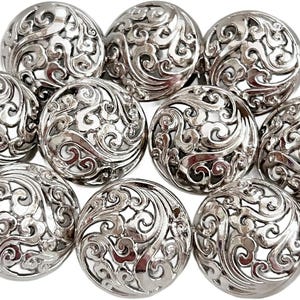 May include: A collection of ten silver-colored, ornate buttons. Each button is round with a detailed, openwork design featuring swirling patterns. The buttons are arranged closely together, showcasing their intricate craftsmanship.