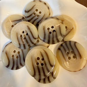 May include: A collection of round, four-hole buttons featuring a cream-colored base with dark brown, vertical, wavy, and striped patterns. Each button has a smooth, glossy finish and a slightly domed surface, arranged in a cluster.