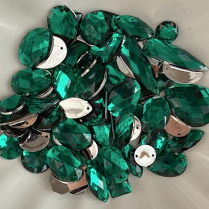 May include: A collection of faceted emerald green gemstones in various shapes, including teardrops, ovals, and rounded forms. Some gems have silver-colored metal backing. The gems are scattered in a white bowl, creating a sparkling display.