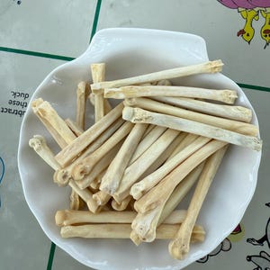 May include: A collection of small, off-white bones are arranged in a white, seashell-shaped dish. The bones have a slightly curved shape and appear to be dried. The dish sits on a patterned surface.