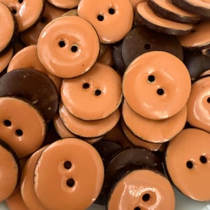 Puede incluir: Una colección de botones redondos en tonos naranja y marrón oscuro. Los botones tienen dos agujeros y un acabado brillante. Los botones están dispersos, mostrando su forma circular y variaciones de color.