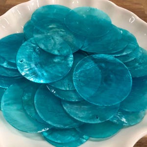 May include: A collection of circular, flat, turquoise-colored shell discs with a pearlescent, iridescent finish. Each thin, translucent disc features a small, singular pre-drilled hole near the edge, suitable for threading or connecting components.