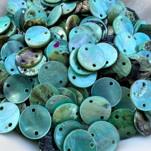 May include: A collection of round, flat abalone shell buttons in shades of turquoise, teal, and green. Each button features a smooth, iridescent surface with natural variations and two small holes drilled near the top for attachment.
