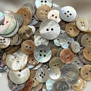 Op de afbeelding: Een verzameling ronde parelmoeren knopen in verschillende maten en tinten iriserend wit, crème, beige en bruin. Elke knoop heeft twee of vier gaten om te naaien, met een glad, gepolijst oppervlak dat de natuurlijke glans van de schelp toont.