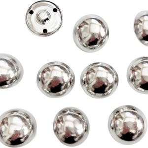 Puede incluir: Una colecci&oacute;n de broches de presi&oacute;n plateados y brillantes. La imagen muestra varias cabezas de broches redondas y abovedadas y una base de broche con cuatro agujeros. Los broches tienen una superficie reflectante, lo que sugiere un material met&aacute;lico.