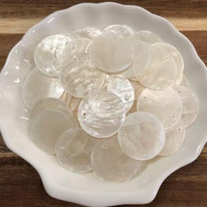 Puede incluir: Una colección de discos circulares de nácar en color blanco roto, cada uno con dos pequeños orificios perforados para su fijación. Estos componentes planos e iridiscentes tienen una superficie lisa y pulida, contenidos en un cuenco blanco festoneado.