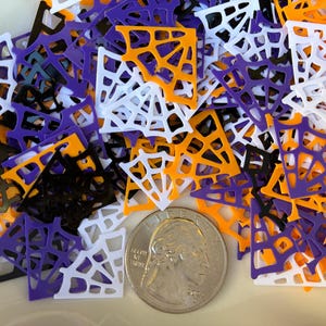 May include: A pile of colorful spiderweb-shaped confetti in orange, purple, white, and black. A U.S. quarter coin is included for size reference. The confetti is likely for Halloween or themed crafts.