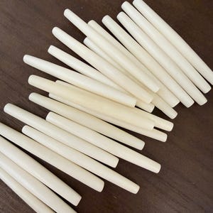 May include: A collection of off-white, cylindrical dowels arranged on a dark brown surface. The dowels are smooth and appear to be made of wood or a similar material. The ends are rounded.