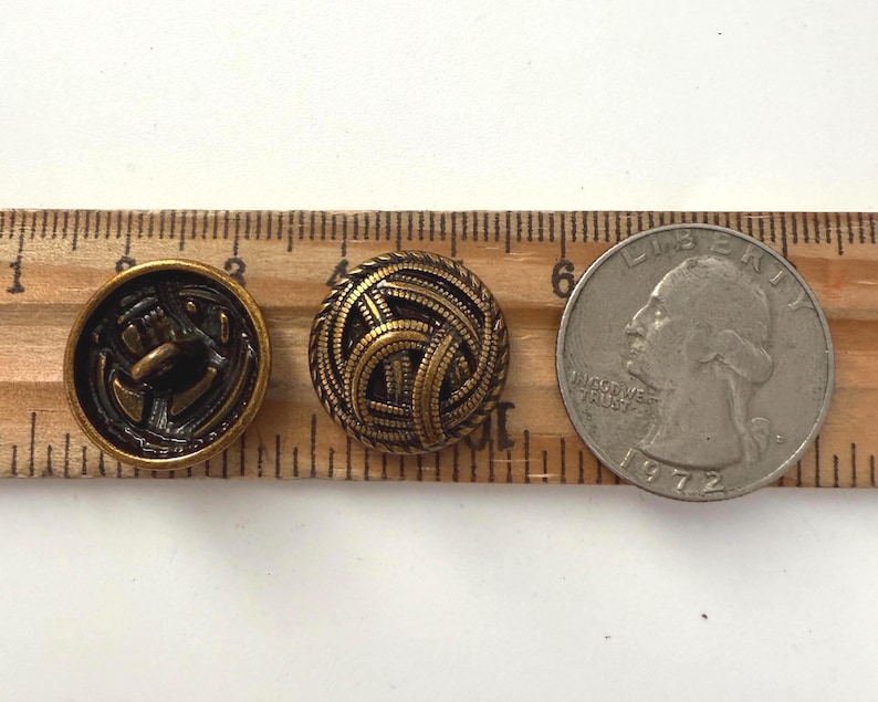 May include: Three antique brass buttons and a quarter coin are displayed on a wooden ruler. The buttons feature intricate designs, including an anchor and a woven pattern. The quarter is dated 1972.