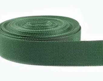 May include: A roll of dark green nylon webbing. The webbing is tightly woven with a ribbed texture, and a portion is unrolled, showing its width. The background is plain white.