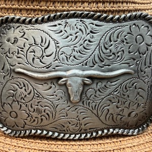 May include: Silver-toned rectangular belt buckle with a longhorn steer design. The buckle features intricate floral and scrollwork patterns and a rope-like border. The buckle is displayed on a straw hat.