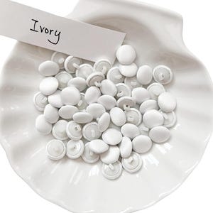 May include: A collection of ivory-colored fabric-covered buttons, displayed in a white, shell-shaped dish. A small white card with the word "Ivory" is placed on top of the buttons. The buttons are round and have a slight sheen.