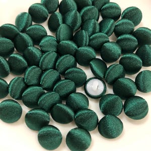 May include: A collection of numerous round buttons covered in a dark emerald green satin fabric. Each button features a smooth, reflective surface with a subtle sheen. One button is flipped over to reveal a white plastic shank on the underside.