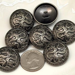 May include: A collection of six round, dark metallic buttons featuring an embossed dragon design with a detailed border. One button is flipped to show the shank back. A 1977 United States quarter coin is included for scale, showing the profile of George Washington.