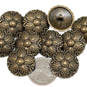 May include: A collection of antique brass-colored buttons with a floral design. The buttons are arranged on a white surface, with a U.S. quarter for size comparison. The buttons have a detailed, embossed pattern.