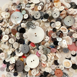 Op de afbeelding: Een verzameling knopen in verschillende maten, vormen en kleuren. De knopen zijn wit, zwart, grijs en beige, met enkele iriserende en parelmoer afwerkingen. Sommige knopen hebben twee gaten, andere vier.