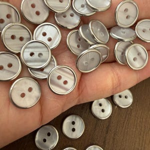 May include: A collection of round, two-hole buttons featuring a pearlescent, off-white surface with subtle gray horizontal banding. Each button is encased in a thin, polished silver-toned metal rim, creating a uniform appearance across the set.