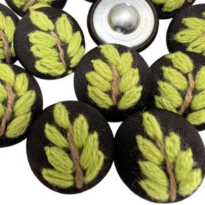 May include: A collection of black fabric buttons with embroidered leaf designs. Each button features a light green leaf pattern on a brown stem. The buttons have a metal backing and are suitable for sewing projects.