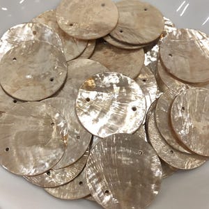 Puede incluir: Una colección de discos circulares de nácar iridiscente, cada uno con dos pequeños orificios perforados. Estos componentes planos y redondos muestran una textura natural brillante en tonos blanco roto y beige con un acabado de superficie pulido y reflectante.