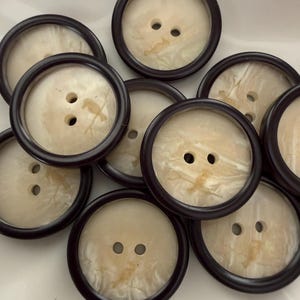 Puede incluir: Una colección de botones grandes y redondos con un centro color crema y un borde marrón oscuro. Cada botón tiene dos agujeros para coser. Los botones tienen un aspecto veteado y son adecuados para proyectos de costura.