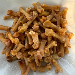 May include: A pile of small, tan-colored bones, possibly animal bones, are clustered together. The bones have a slightly curved shape with rounded ends, and some have a darker, almost burnt appearance. The background is a white, translucent surface.