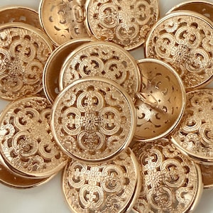 May include: A pile of gold-coloured, decorative buttons. Each button features an intricate, openwork design with a central cross motif and swirling patterns. The buttons are round and slightly domed, suggesting a vintage or decorative style.