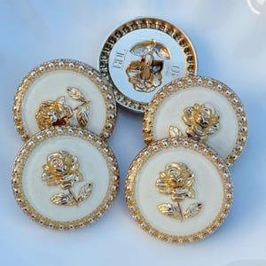 May include: A collection of decorative buttons with a cream-colored center and gold-colored rose and leaf designs. The buttons have a gold-colored beaded border and a silver-colored back. The buttons are round.