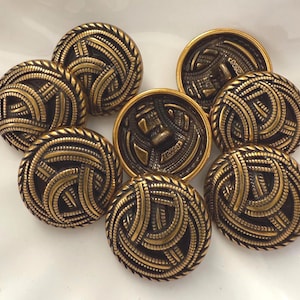May include: A collection of eight antique brass buttons. The buttons have a detailed design of interwoven lines and a rope-like border. The buttons are round and have a dark brass color with gold accents.