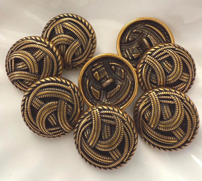 May include: A collection of eight antique brass buttons. The buttons have a detailed design of interwoven lines and a rope-like border. The buttons are round and have a dark brass color with gold accents.