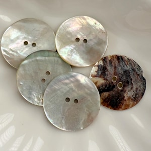 May include: A collection of five round, iridescent mother-of-pearl buttons. Each button has two holes for sewing. The buttons vary in color, with shades of white, silver, and brown. The buttons are arranged on a white surface.