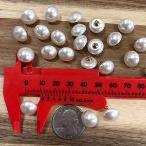 May include: A collection of round, white, pearl-like buttons with a smooth, glossy finish. A bright red plastic caliper is positioned over one button, showing a diameter of approximately 0.5 inches, alongside a silver US quarter coin for scale.