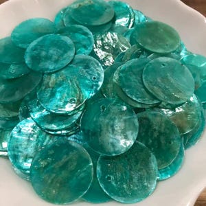 Peut inclure: Une collection de disques en coquillage circulaires de couleur sarcelle avec une finition irisée naturelle. Chaque pièce plate et ronde présente un petit trou pré-percé près du bord, mettant en valeur la texture organique et la surface nacrée.