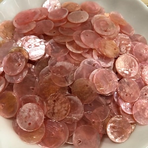 Puede incluir: Una colección de discos circulares de concha de color rosa claro con un acabado nacarado. Cada disco plano y delgado presenta un pequeño orificio cerca del borde, mostrando variaciones naturales de color y textura en el conjunto.