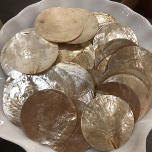 Peut inclure: Une collection de disques circulaires et plats en nacre blanc cassé, présentant un éclat irisé naturel et une texture lisse. Ces pièces de coquillage rondes sont empilées et superposées, affichant des tons variés de brun clair et de crème.