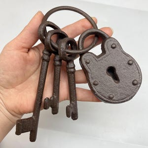 May include: A set of antique-style keys and a padlock. The keys are attached to a large ring, and the padlock has a keyhole. The metal has a rustic, brown finish, suggesting an aged appearance. The keys have detailed designs.