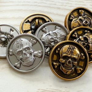 Può includere: Una collezione di bottoni metallici con disegni di teschi e ossa incrociate. I bottoni presentano finiture metalliche varie, tra cui argento e oro. I dettagli del teschio sono ben visibili su ogni bottone.