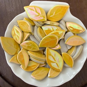 May include: A white, shell-shaped dish filled with decorative ceramic leaf tiles. The tiles are primarily yellow with iridescent highlights, and some are a light gray color. The leaves are arranged in a random pattern.