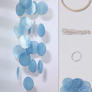 May include: A hanging mobile featuring multiple translucent, circular, light blue shell discs suspended from a circular wooden frame by thin white strings. The set includes a wooden hoop, a coil of white string, and a pile of loose blue shell discs.